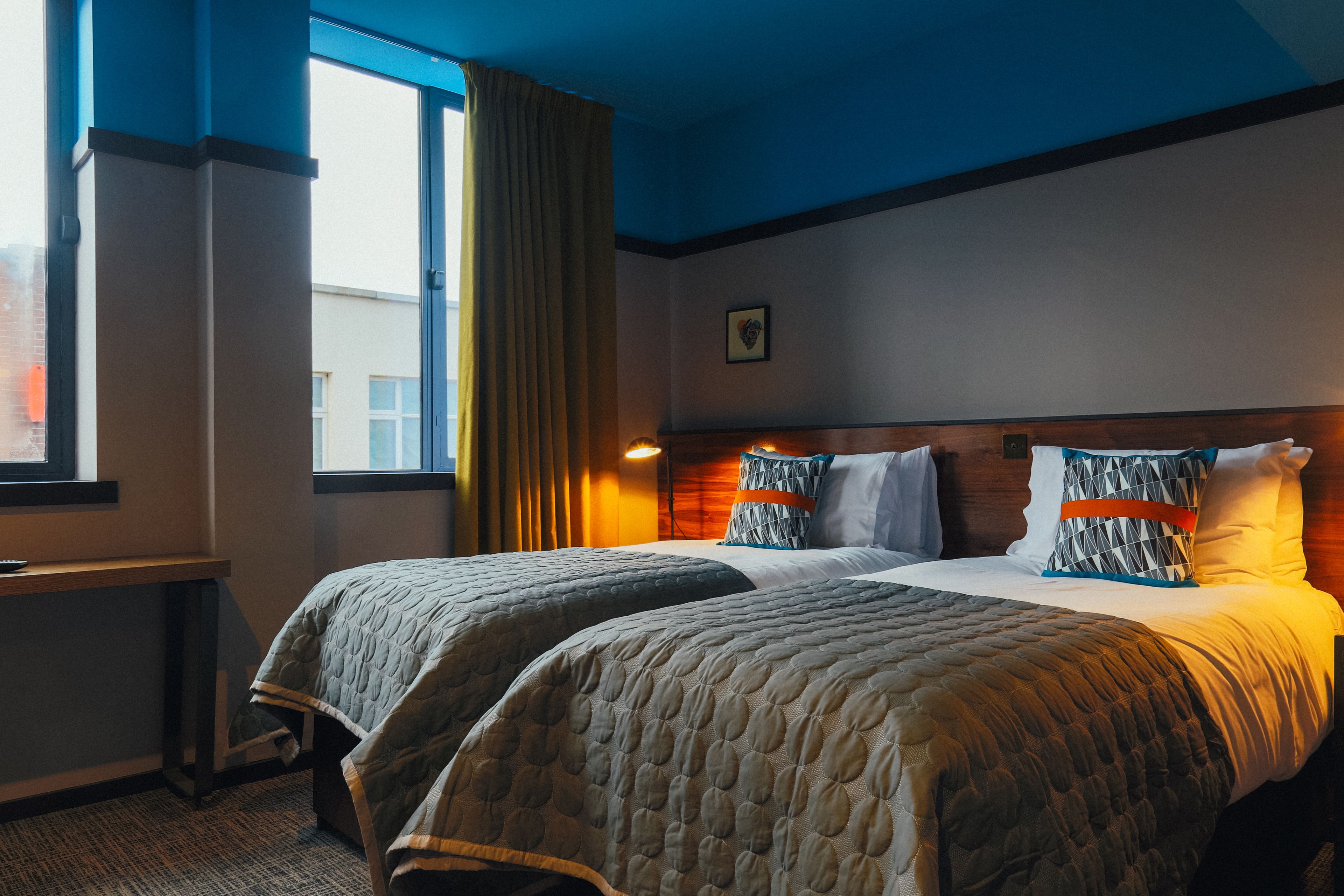 A hotel room with two beds, a window with curtains, a bedside table, and a wall-mounted picture.