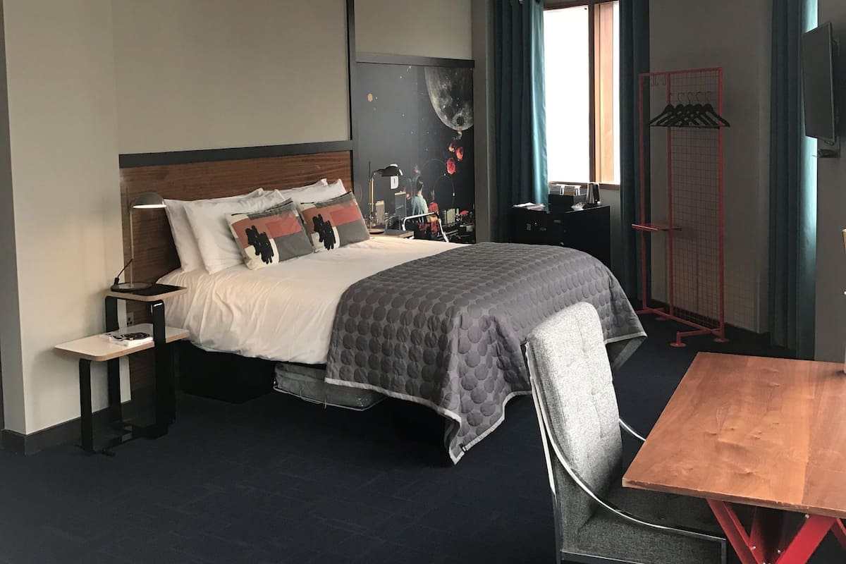 A modern hotel room with a bed, bedside table, chair, and a small table.
