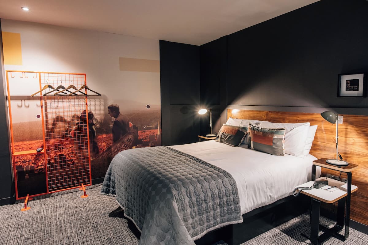 A modern hotel room with a large bed, a wooden headboard, a bedside table with a lamp, and a wall-mounted TV.