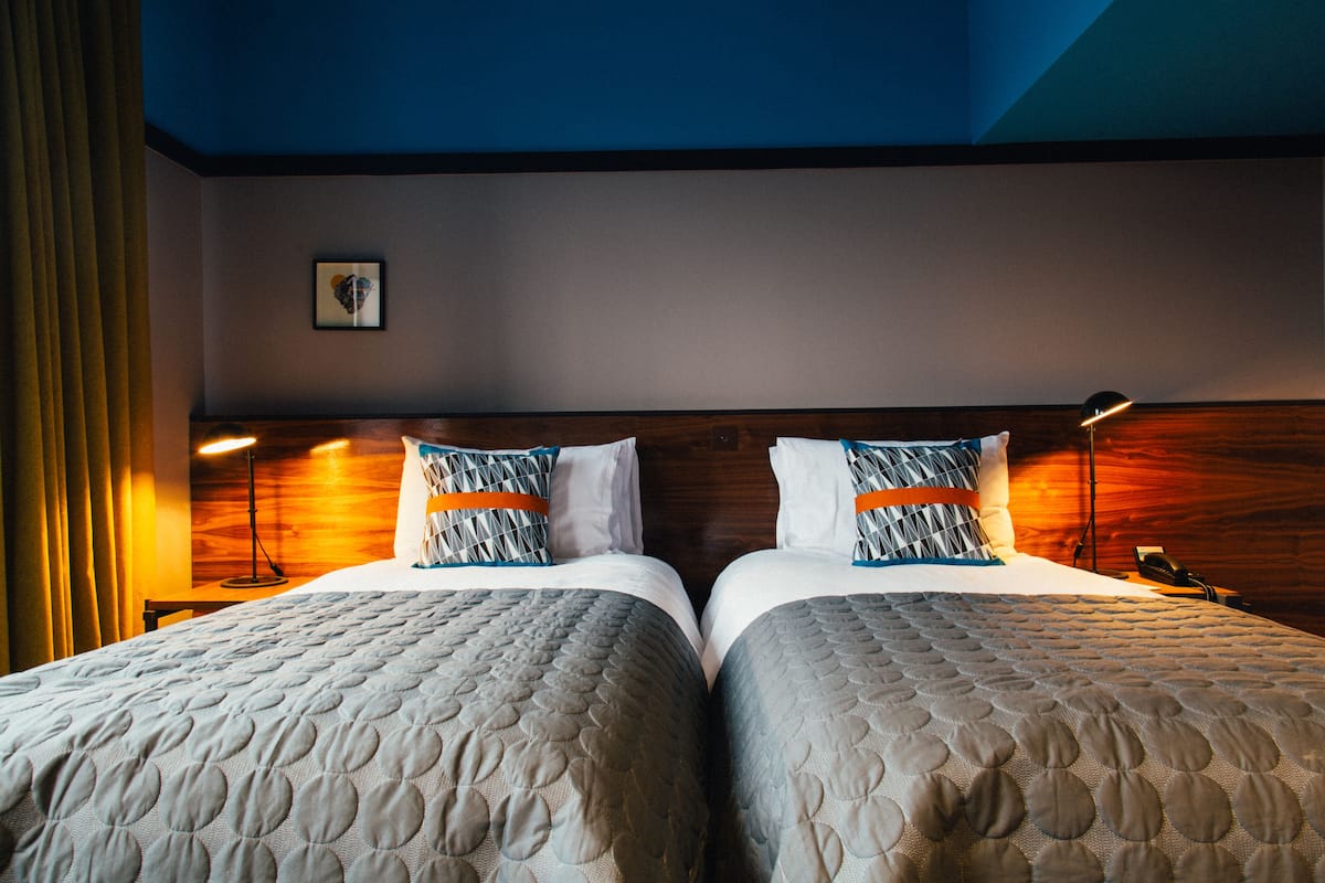 A hotel room with two beds, a wooden headboard, a framed picture on the wall, and a nightstand with a lamp.