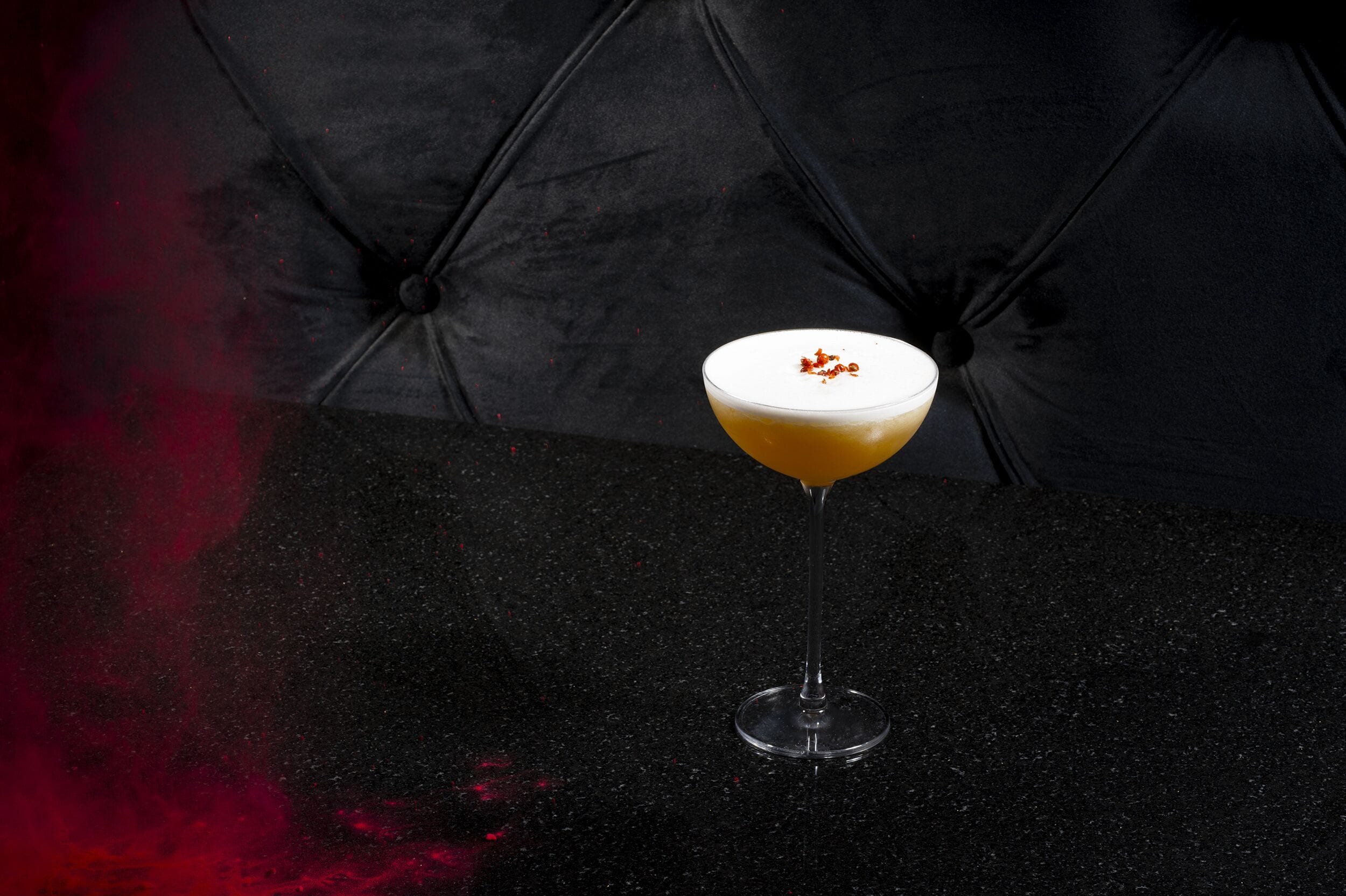 A cocktail in a martini glass on a black table with a dark, textured background.