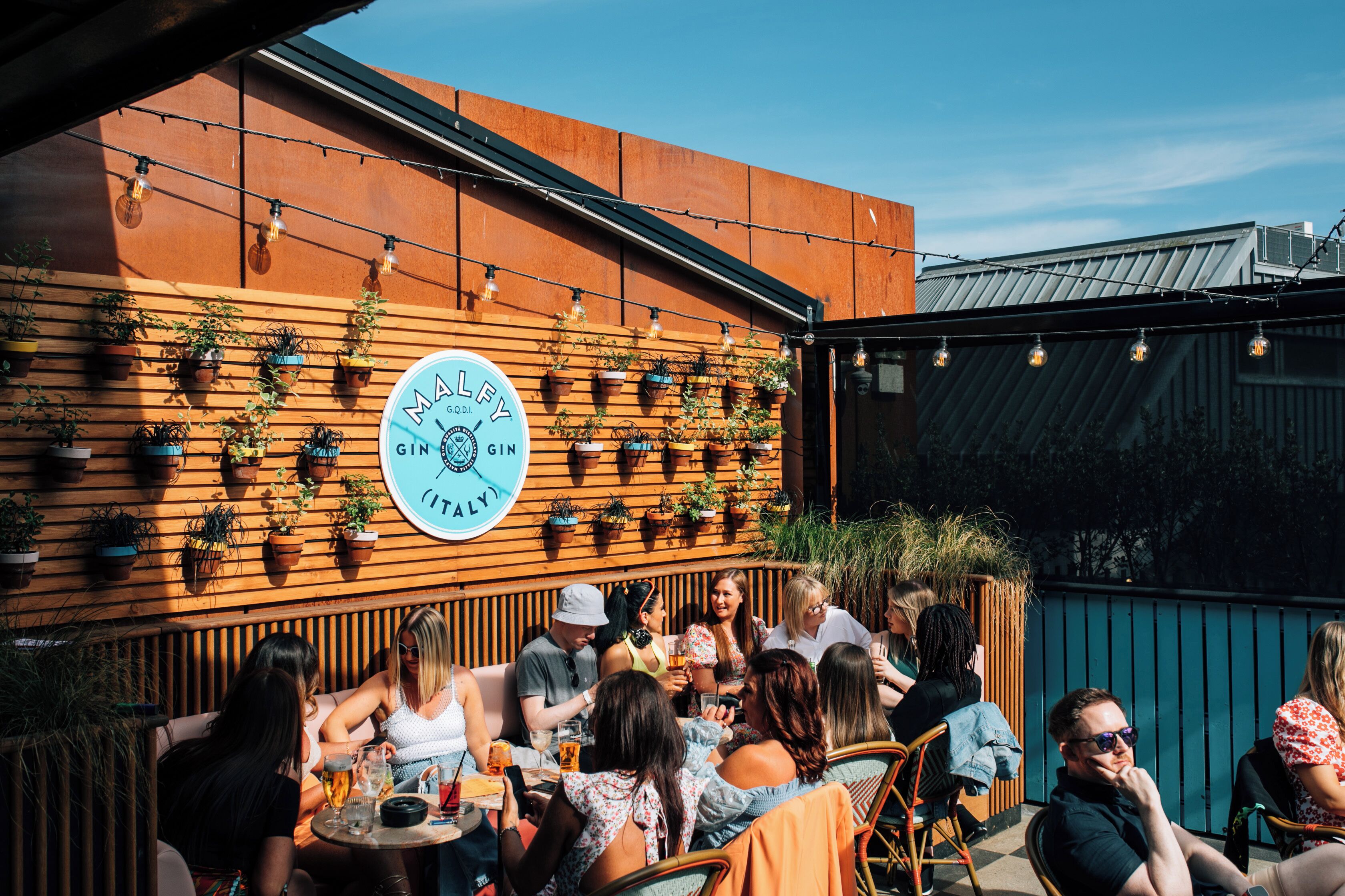 A group of people seated at tables outdoors, with a wooden wall adorned with potted plants and a large circular sign.