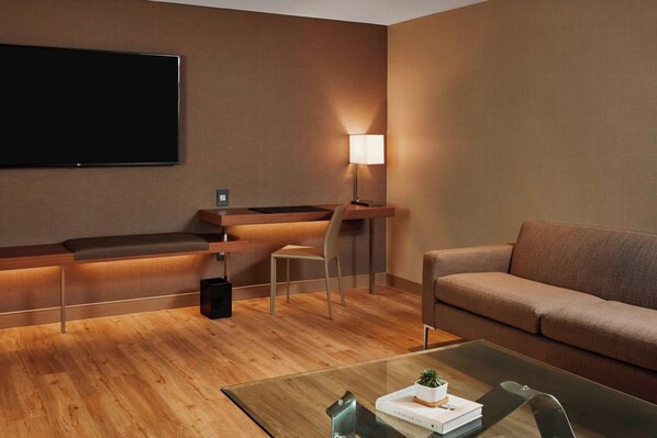 Una habitación de hotel moderna con una televisión de pantalla plana, un escritorio con lámpara, un sofá y una mesa de centro de vidrio con un libro y una planta.