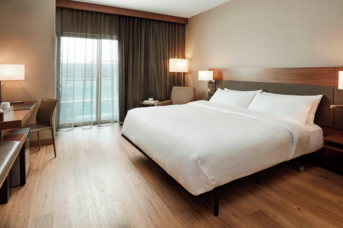 Ein Hotelzimmer mit einem großen Bett, einem Schreibtisch, einem Sessel und Blick ins Freie durch ein Fenster.