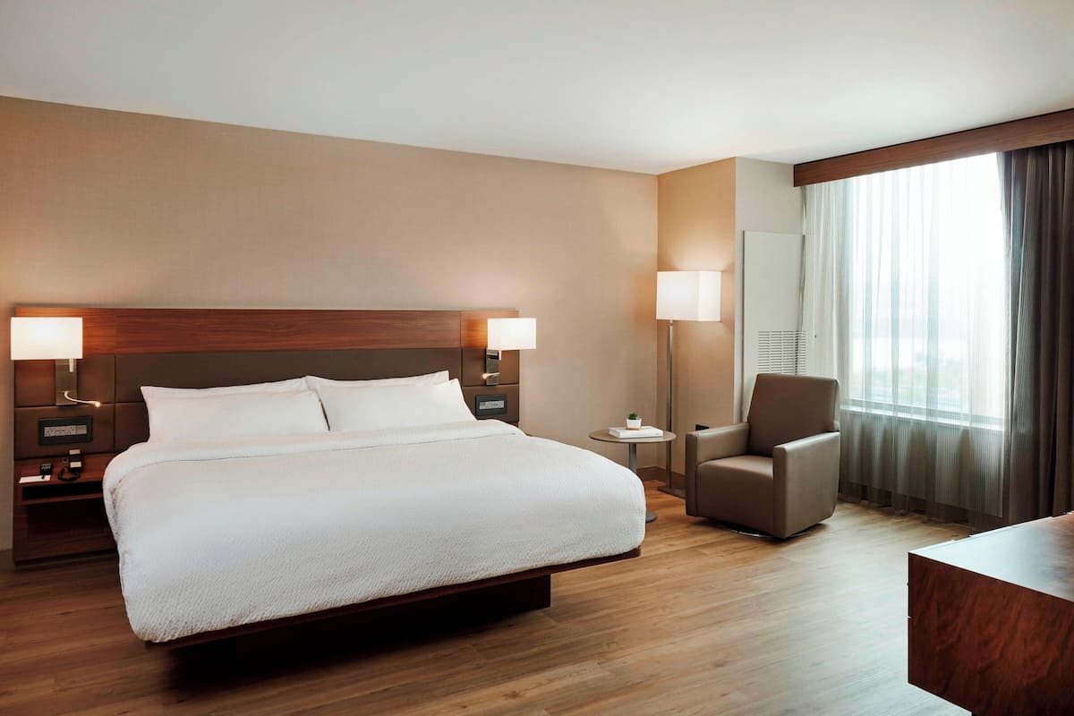 Ein Hotelzimmer mit einem großen Bett, zwei Nachttischlampen, einem Sessel, einem kleinen Tisch und einem großen Fenster mit Vorhängen.