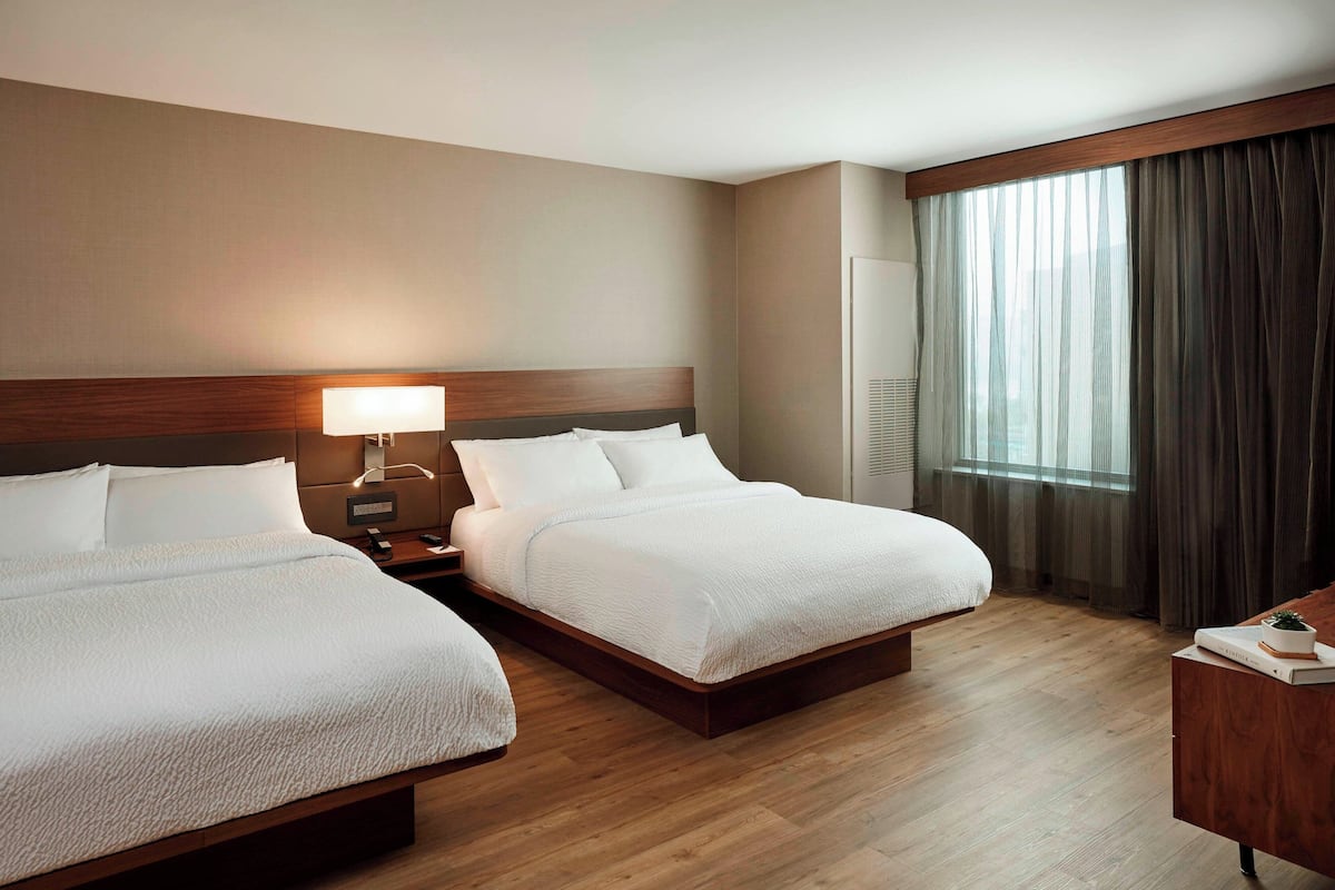 Ein Hotelzimmer mit zwei Betten, einem Nachttisch mit Lampe, einem Holz-Kopfteil, einem großen Fenster mit Vorhängen und einem Schreibtisch mit Pflanze.