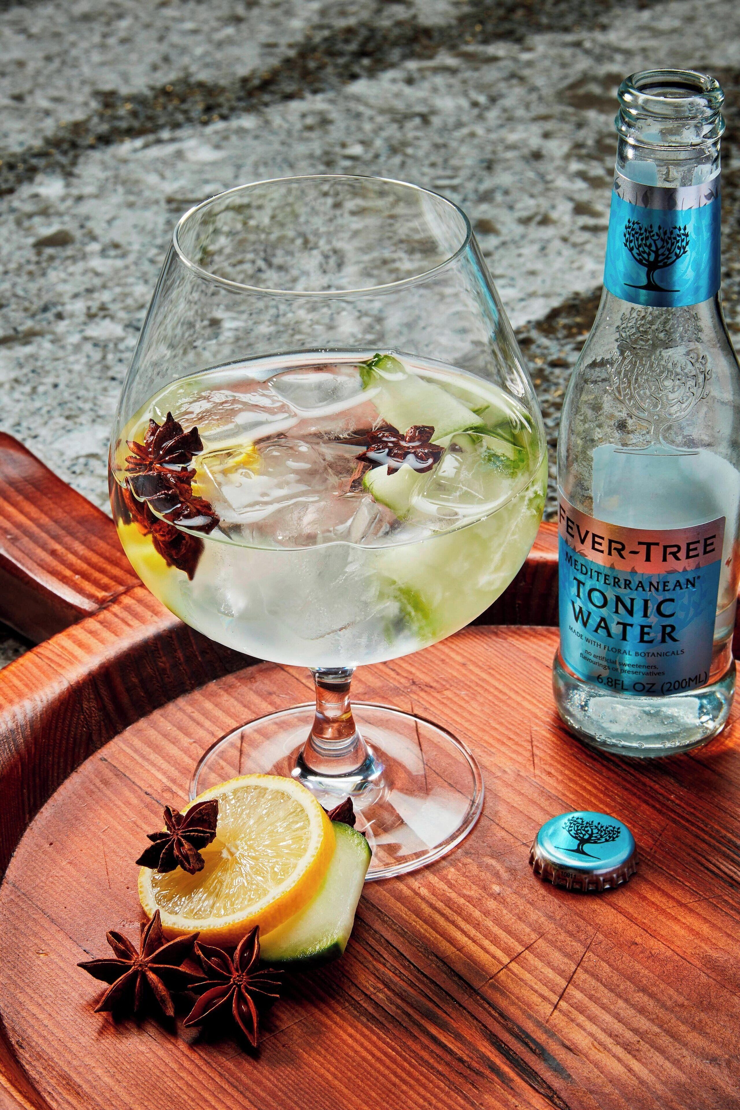 Un vaso de agua tónica con limón y estrella de anís sobre una bandeja de madera, junto a una botella que dice 'Fever-Tree Mediterranean Tonic Water'.