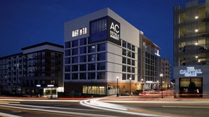 A modern hotel with a prominent 'AC Hotels Marriott' sign, a well-lit facade, and a visible street with light trails indicating traffic. - AC Hotel Raleigh North Hills (Raleigh)