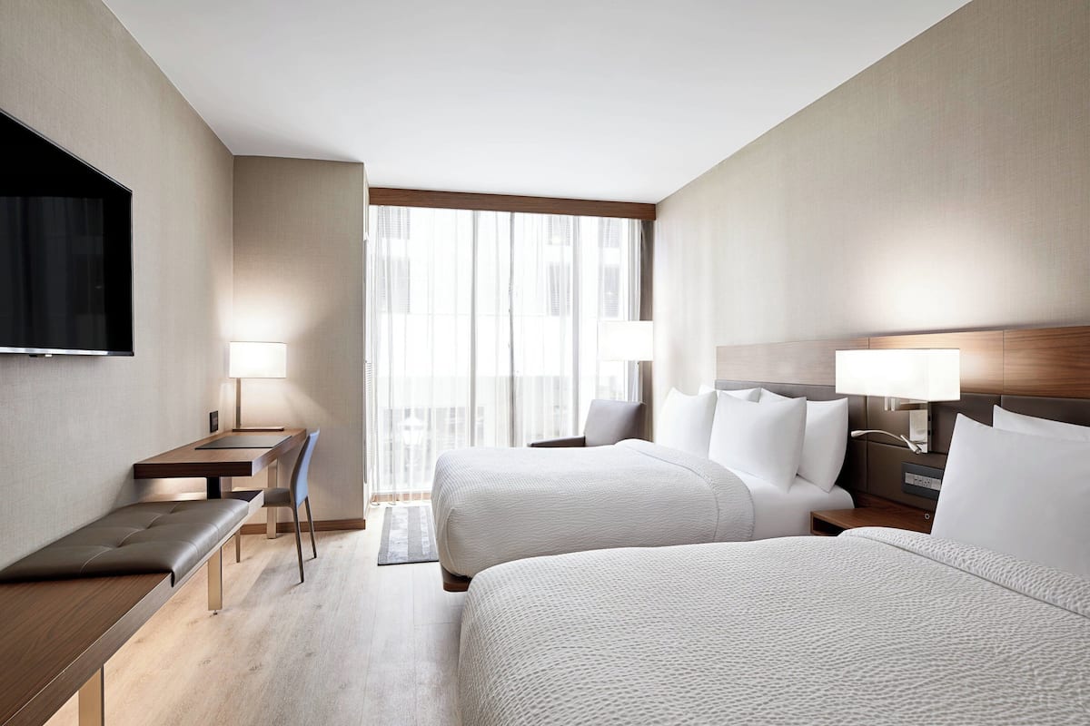 A hotel room with two beds, a desk, a chair, a TV, and a large window with curtains.