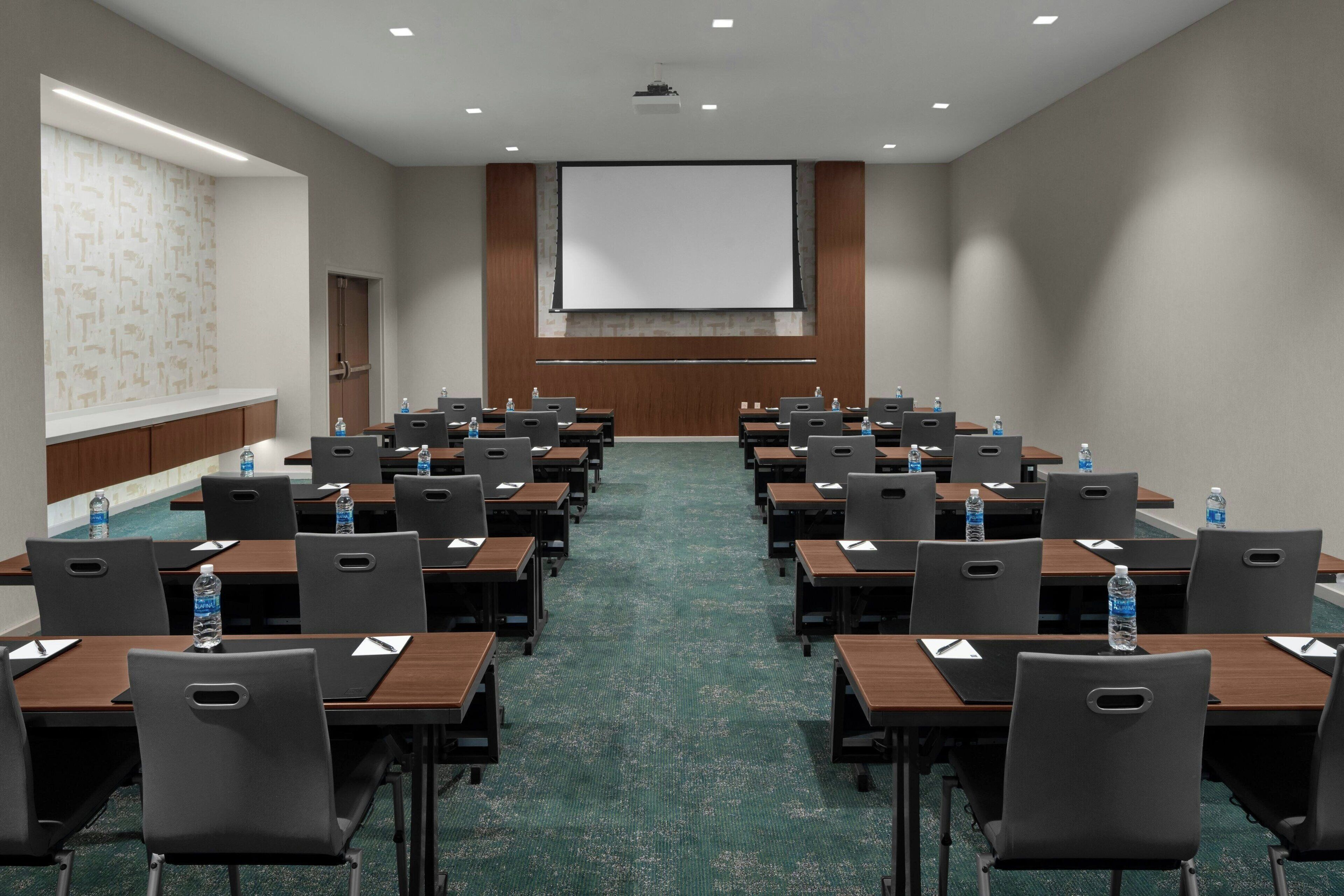 A conference room with rows of tables and chairs, a projector screen, and a water dispenser.