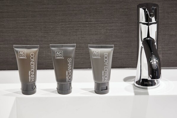 Tres productos de tocador en tubos grises con la etiqueta 'AC Hotels MARBETA FRESH NEROLI' y un grifo cromado con la marca 'DELTA' visible.