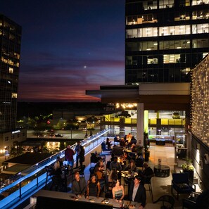 Un bar en la azotea con mesas al aire libre, rodeado de edificios modernos y vistas de la ciudad al atardecer.