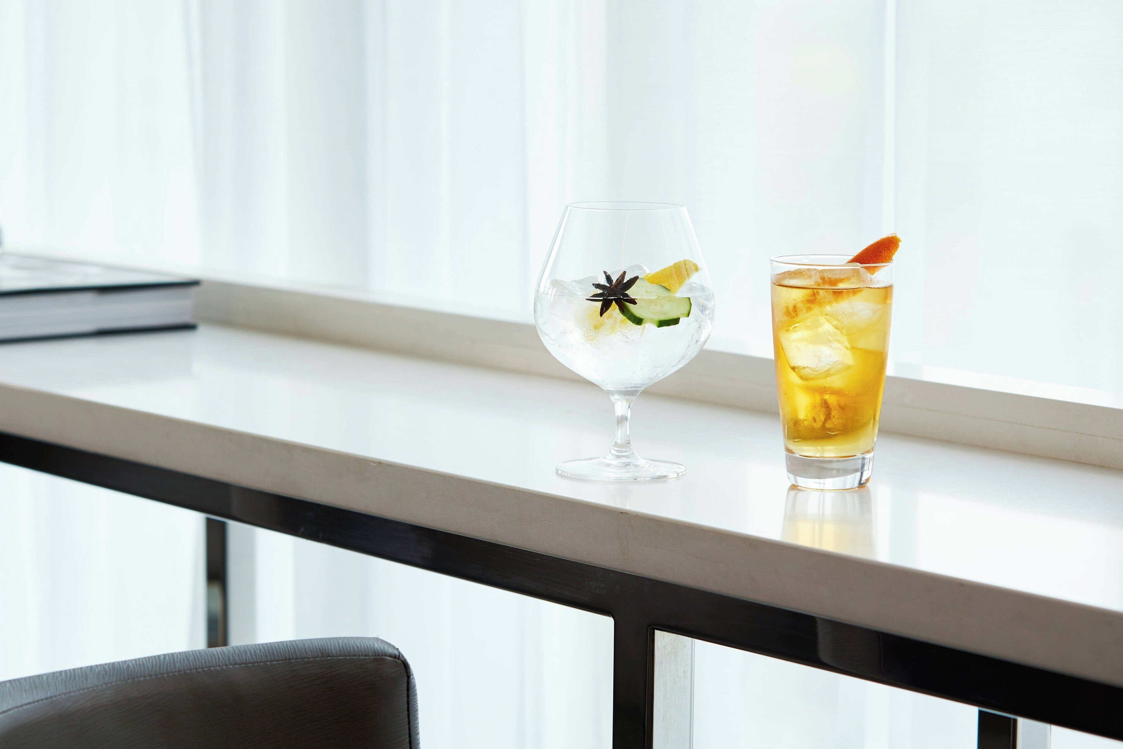 Two glasses of beverages on a table near a window with sheer curtains.