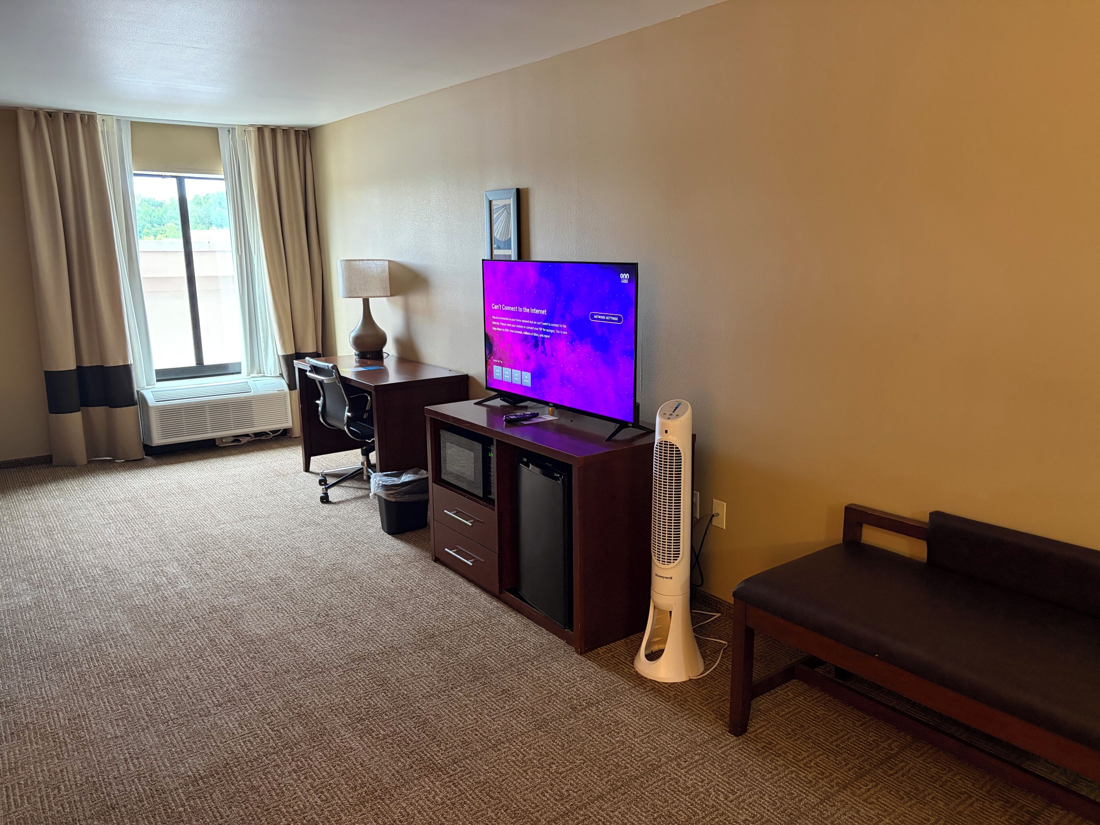 A hotel room with a desk, chair, television, and a standing fan.