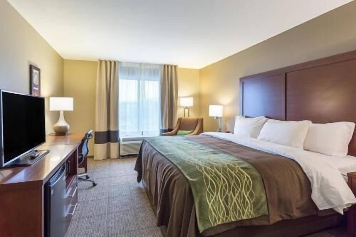 A hotel room with a bed, a desk with a television, two bedside lamps, a chair, and a window with curtains.
