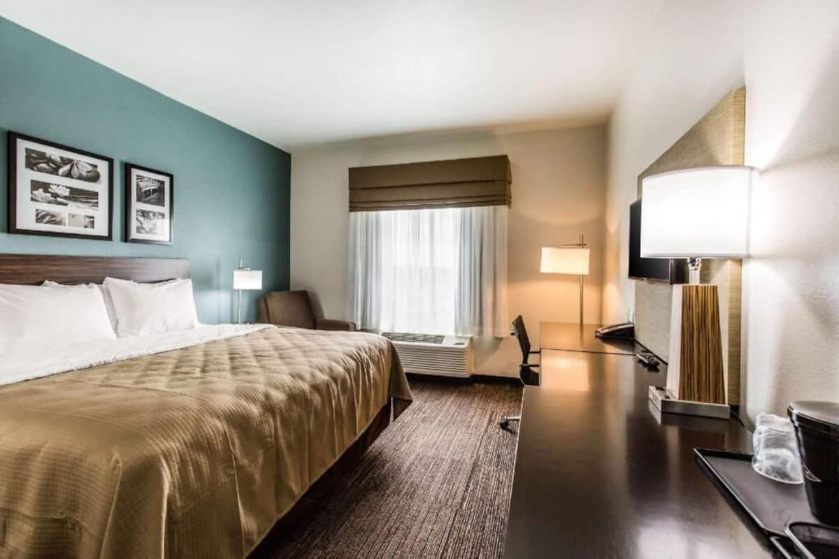 A hotel room with a bed, a desk with a lamp, a chair, a window with curtains, and framed pictures on the wall.