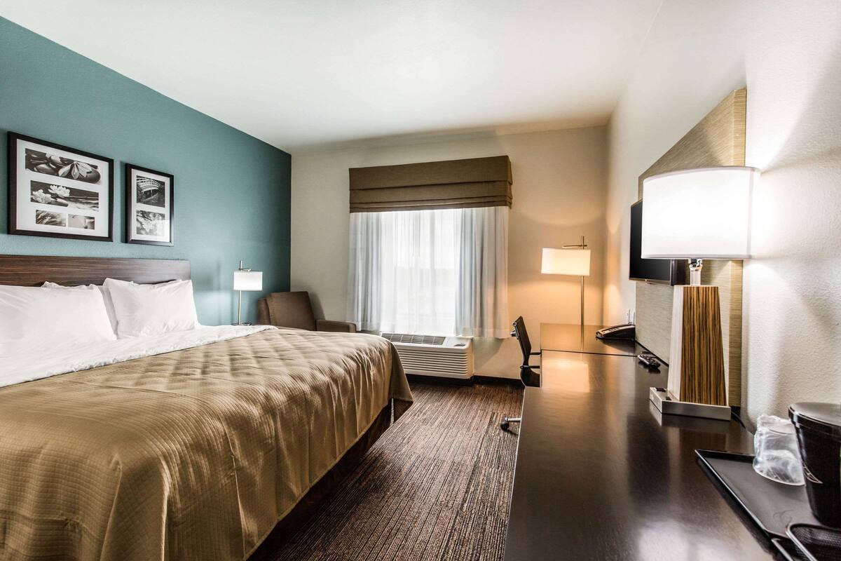 A hotel room with a large bed, a desk with a lamp, a chair, and a window with curtains.