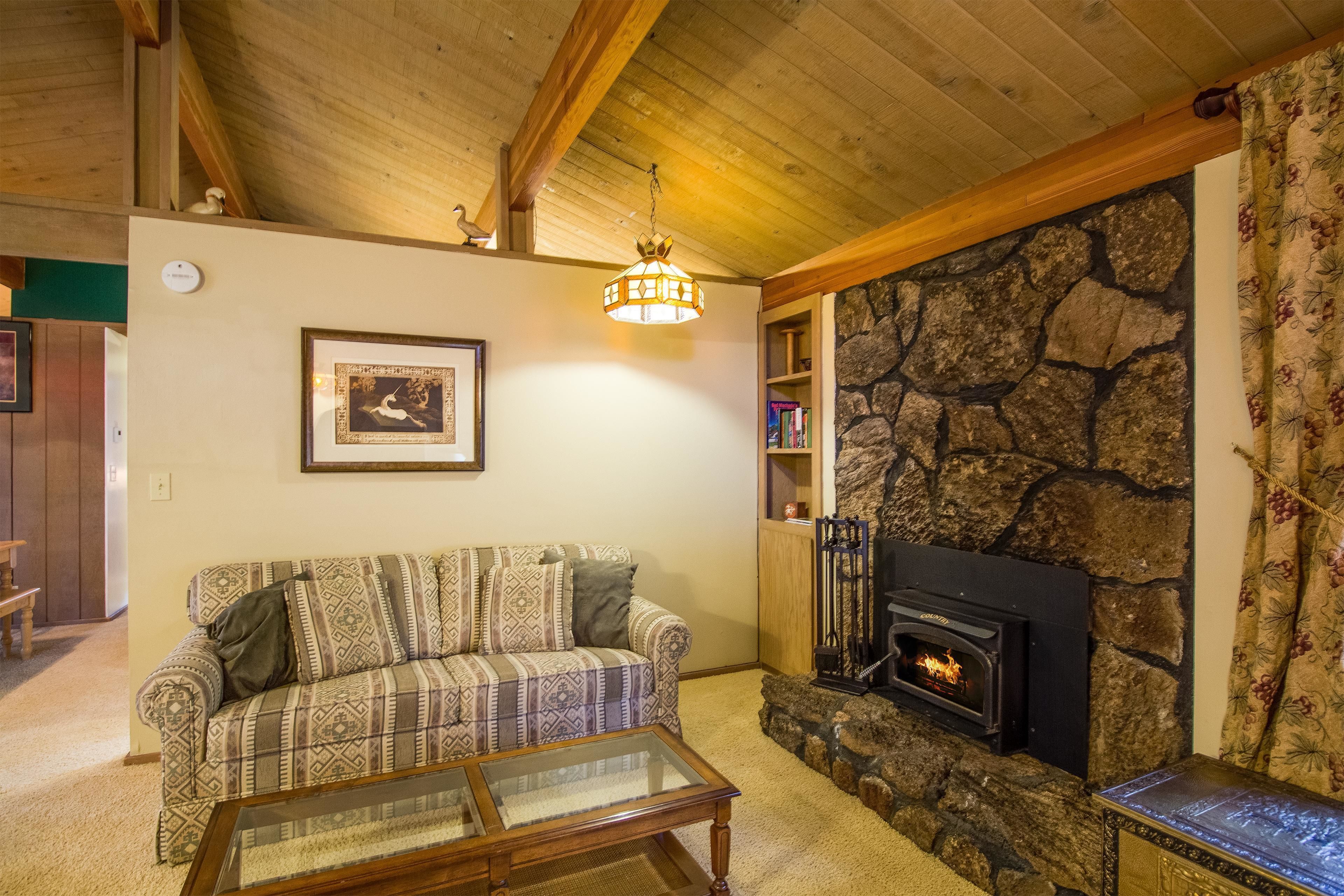 A cozy living room with a patterned sofa, a glass coffee table, a stone fireplace, and a wooden ceiling.