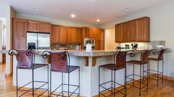 Una cocina con armarios de madera, un refrigerador de acero inoxidable, un microondas y una encimera de granito. Hay cuatro taburetes de bar con estructura metálica y asientos de cuero.