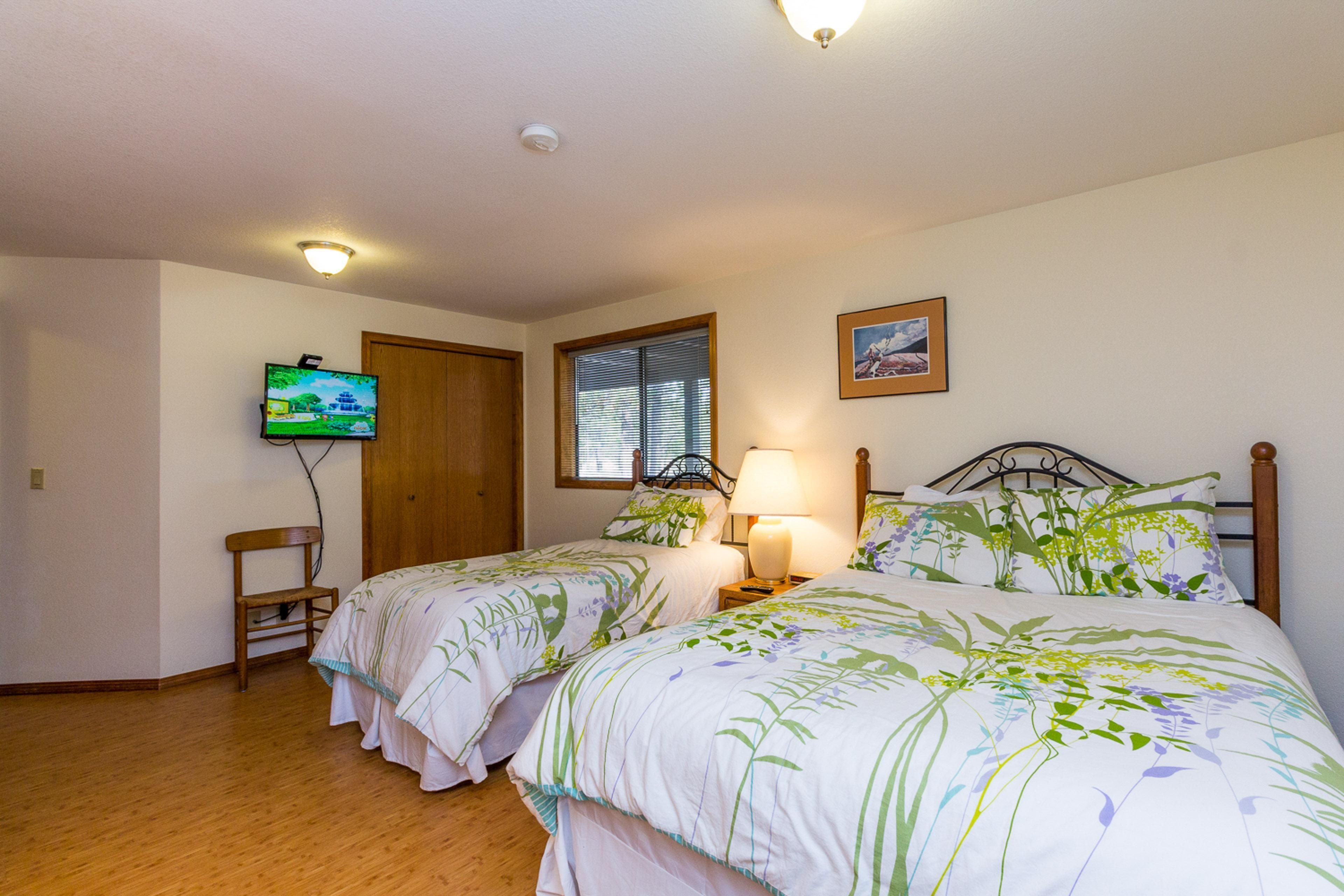 Ein Schlafzimmer mit zwei Einzelbetten, einem an der Wand montierten Fernseher, einem Holzstuhl, einem Nachttisch mit Lampe und einem Fenster mit Jalousien.