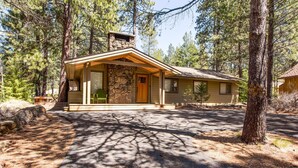 A rustic cabin with a stone chimney, a covered porch, and a driveway leading to the entrance. - Grizzly 5 (Bend)