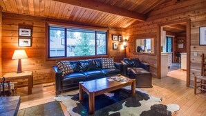 A cozy log cabin interior with a wooden ceiling, a large window, a leather sofa, a coffee table, and a cowhide rug. - Grizzly 5 (Bend)