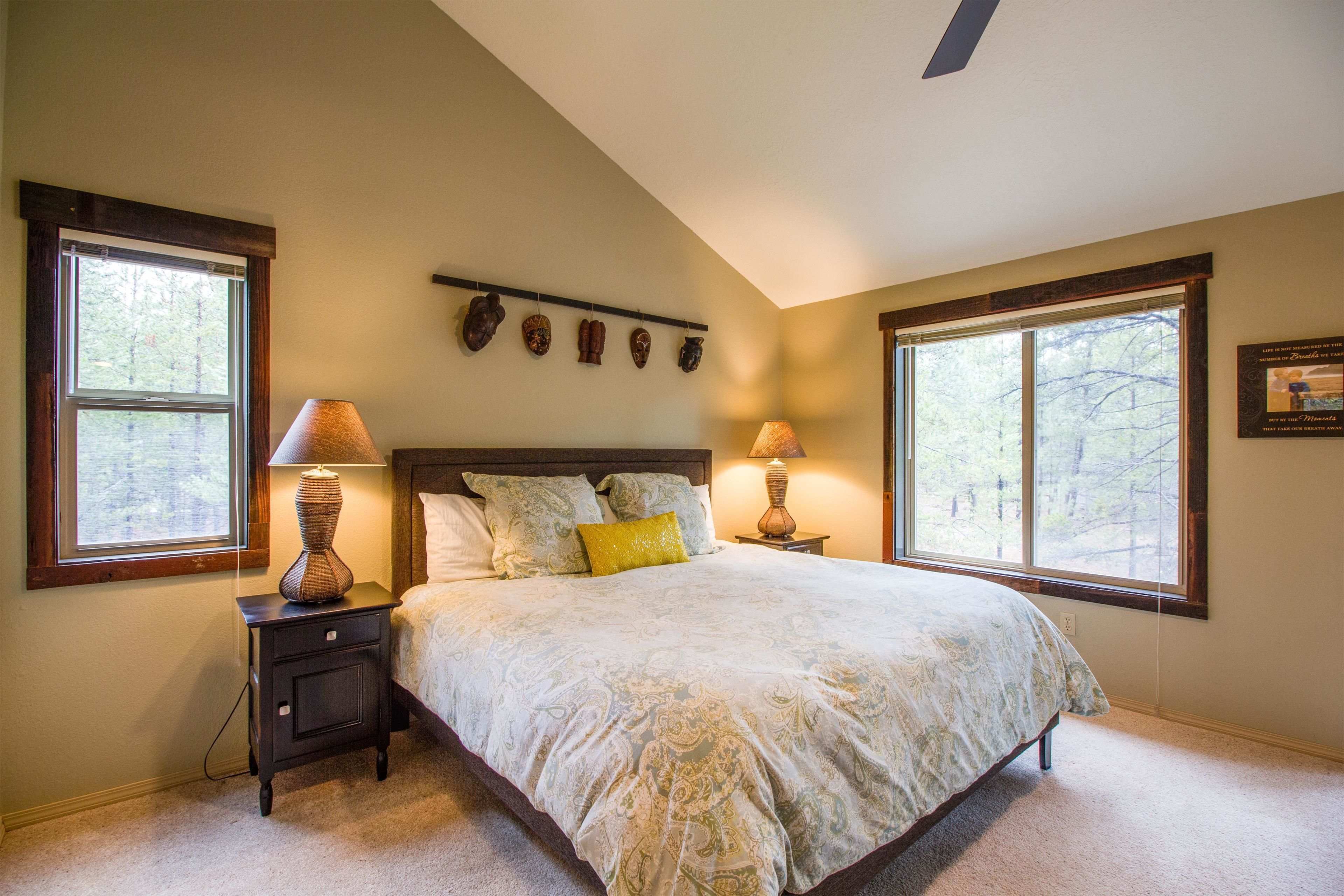 A bedroom with a large bed, two bedside tables, a ceiling fan, and a window with a view of trees.