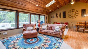 A spacious living room with a large wooden ceiling, a patterned rug, a brown leather sofa, and a dining area with wooden furniture. - Hoodoo 5 (Bend)