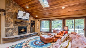 A cozy living room with a stone fireplace, leather sofa, and large windows offering a view of the outdoors. - Hoodoo 5 (Bend)