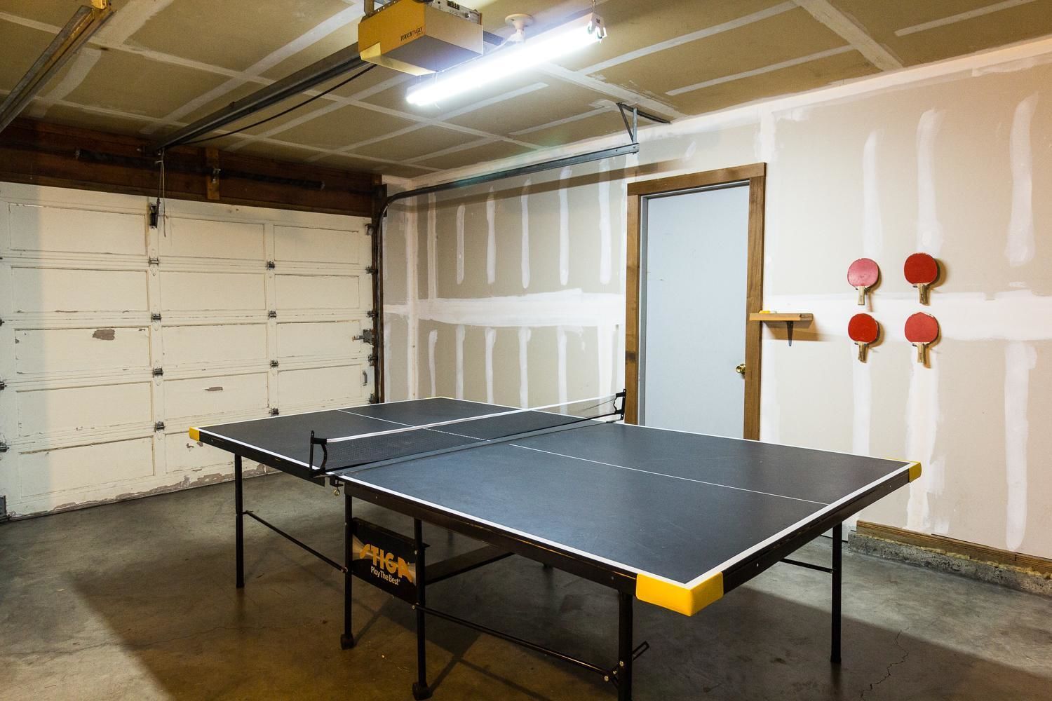 A ping pong table in a garage with a white door and a wall with red ping pong paddles.