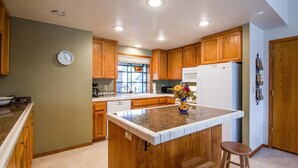 A kitchen with wooden cabinets, a central island with stools, a clock on the wall, and a window with blinds.