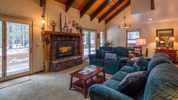 A cozy living room with a stone fireplace, a ceiling fan, and a large window overlooking a snowy landscape. - Leisure 4 (Bend)