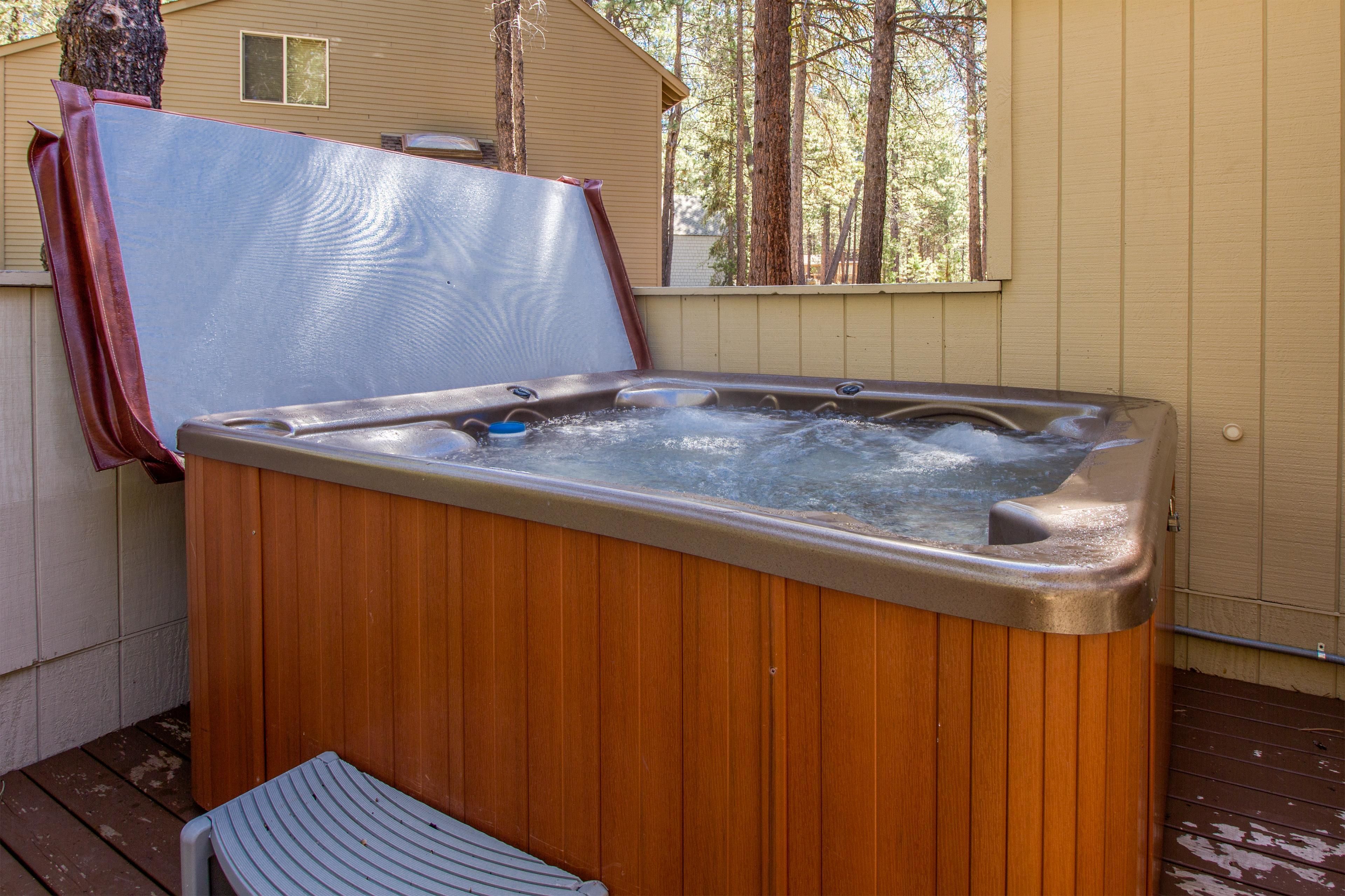 A wooden hot tub with a bench, surrounded by a wooden deck and a house with a window and trees in the background.
