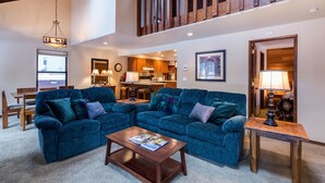 A spacious living room with a large blue sectional sofa, a wooden coffee table, and a dining area with wooden chairs. A kitchen is visible in the background, and there is a staircase leading to an upper level. - Leisure 4 (Bend)