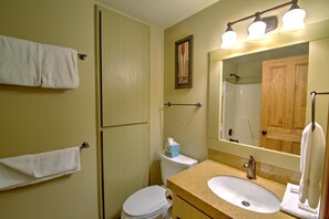 Un baño con inodoro, lavabo y espejo. Hay un perchero con toallas, un cuadro en la pared y una barra para cortina de ducha.