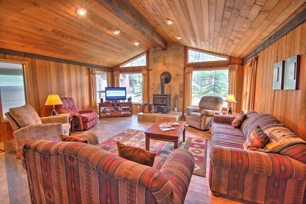 Una sala de estar acogedora con paredes y techo de madera, una chimenea de piedra y ventanas amplias que ofrecen vistas al exterior.