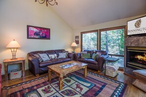 A cozy living room with a leather sofa, a coffee table, and a fireplace. There is a large window with a view of trees. - Sparks 5 (Bend)