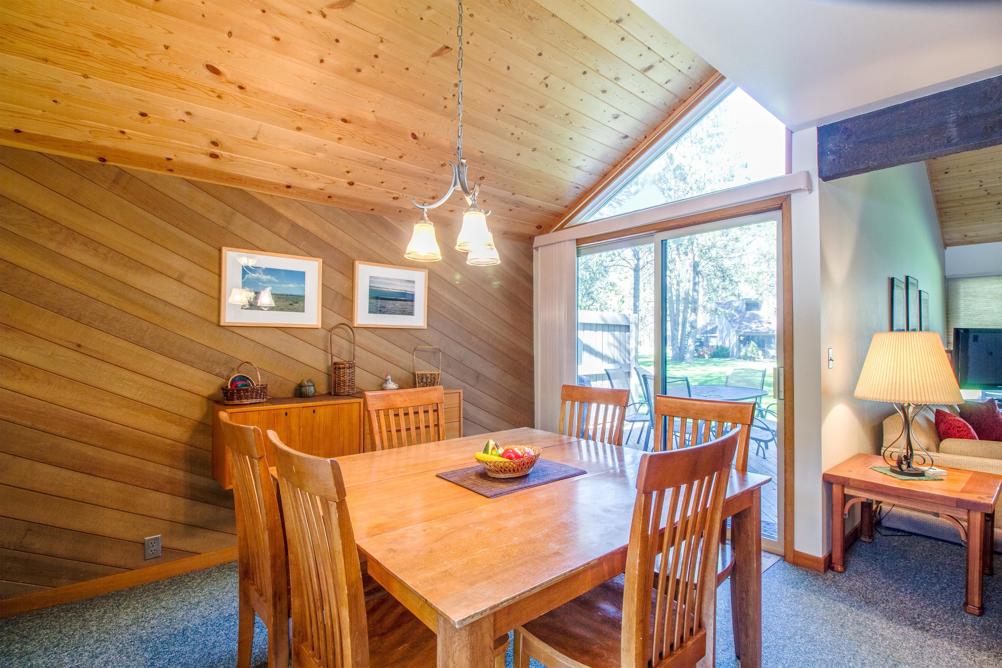 A wooden dining area with a table, chairs, and a bowl of fruit. There are two framed pictures on the wall, a lamp on a side table, and a window with a view of the outdoors.
