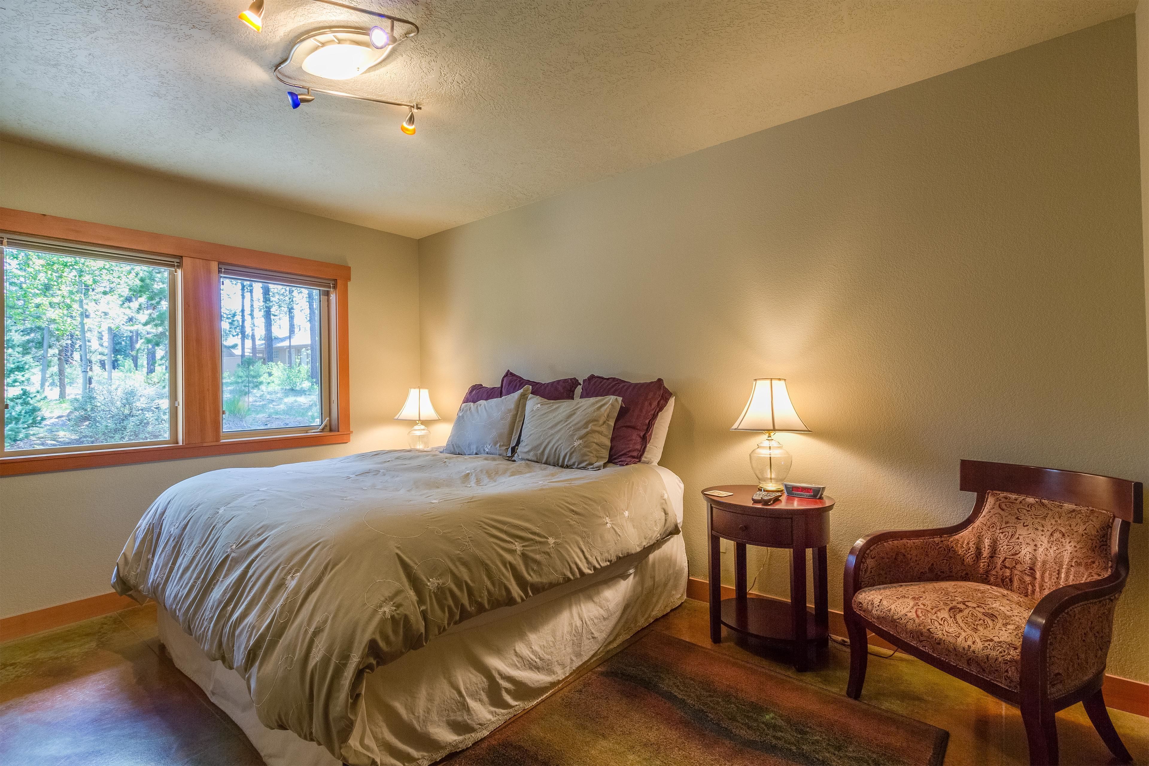 A bedroom with a large bed, two bedside lamps, a chair, and a window with a view of trees.