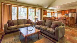 A spacious living room with two sofas and a coffee table, a kitchen with wooden cabinets and a refrigerator, and large windows offering a view of the outdoors. - Mt Rose 9 (Bend)