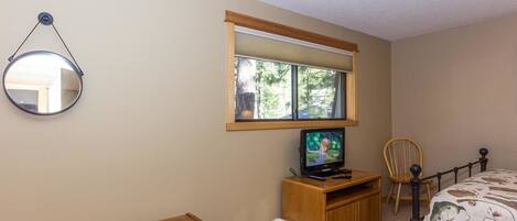 A bedroom with a bed, a TV on a wooden stand, a window with a view of trees, a mirror on the wall, and a small wooden dresser.