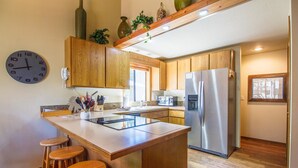A modern kitchen with wooden cabinets, a stainless steel refrigerator, a built-in island with stools, and a large wall clock. - Nine Iron 10 (Bend)
