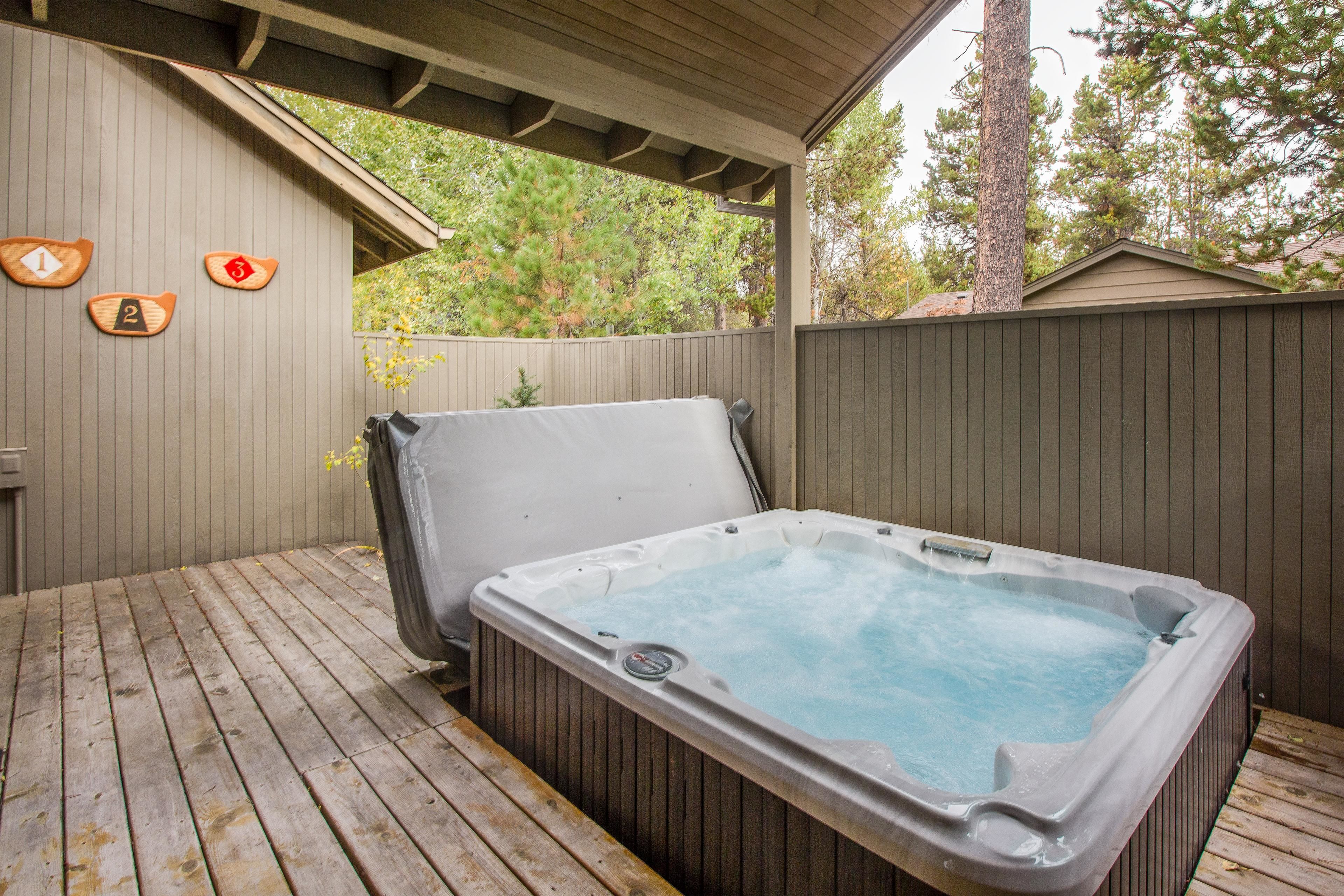 A hot tub on a wooden deck with a covered area, wooden fence, and trees in the background.