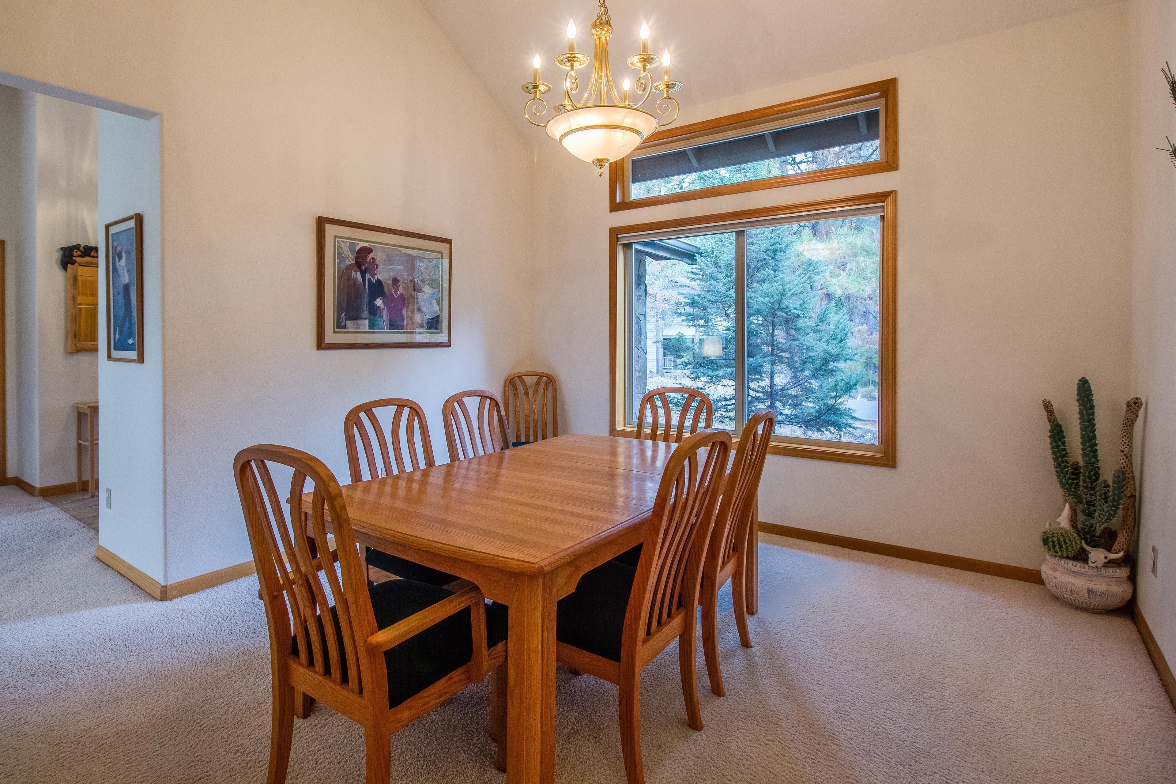A dining room with a wooden table and chairs, a chandelier, framed pictures on the wall, and a large window with a view of trees.