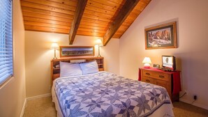 A bedroom with a wooden ceiling, a bed with a patterned bedspread, two bedside lamps, a nightstand with a lamp, a framed picture on the wall, and a dresser with a red cloth. - Elk Lane 7 (Bend)