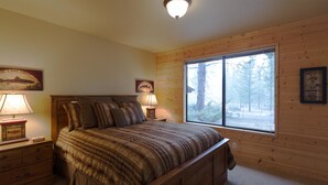 A bedroom with a wooden bed, two bedside tables with lamps, a large window with a view of trees, and framed pictures on the walls. - Pioneer 7 (Bend)