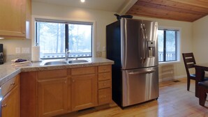 A kitchen with wooden cabinets, a stainless steel refrigerator, and a window with a view of trees. - Pioneer 7 (Bend)