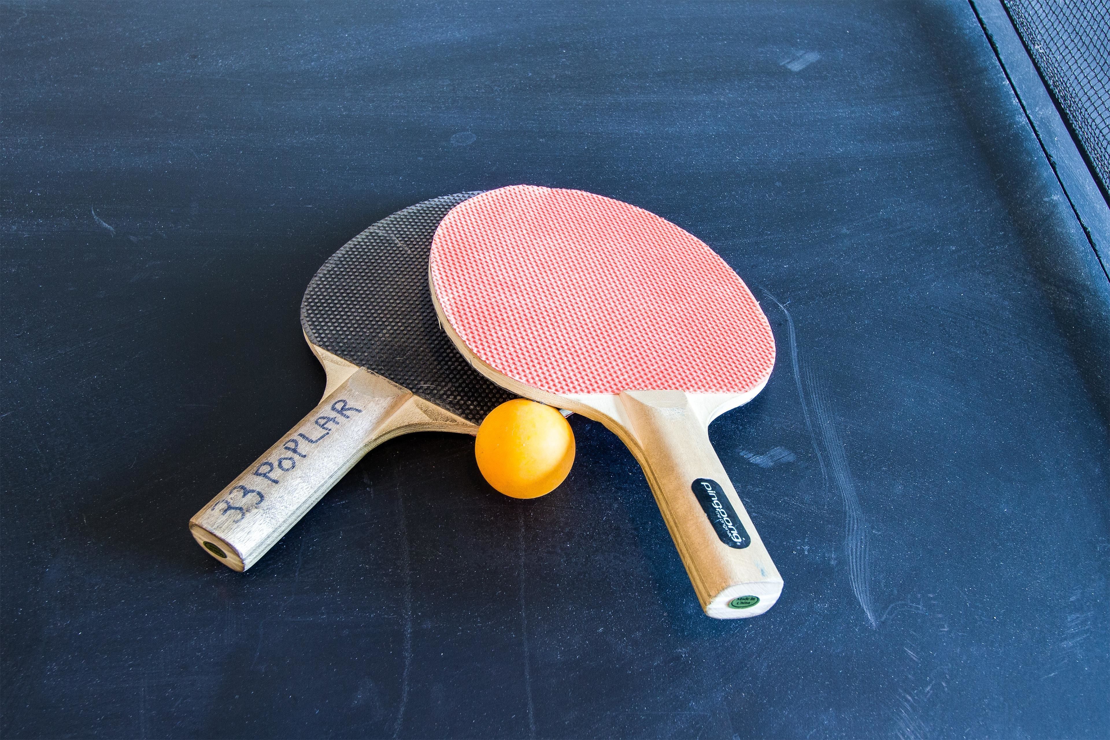 Two table tennis paddles with a yellow ball between them on a dark surface.