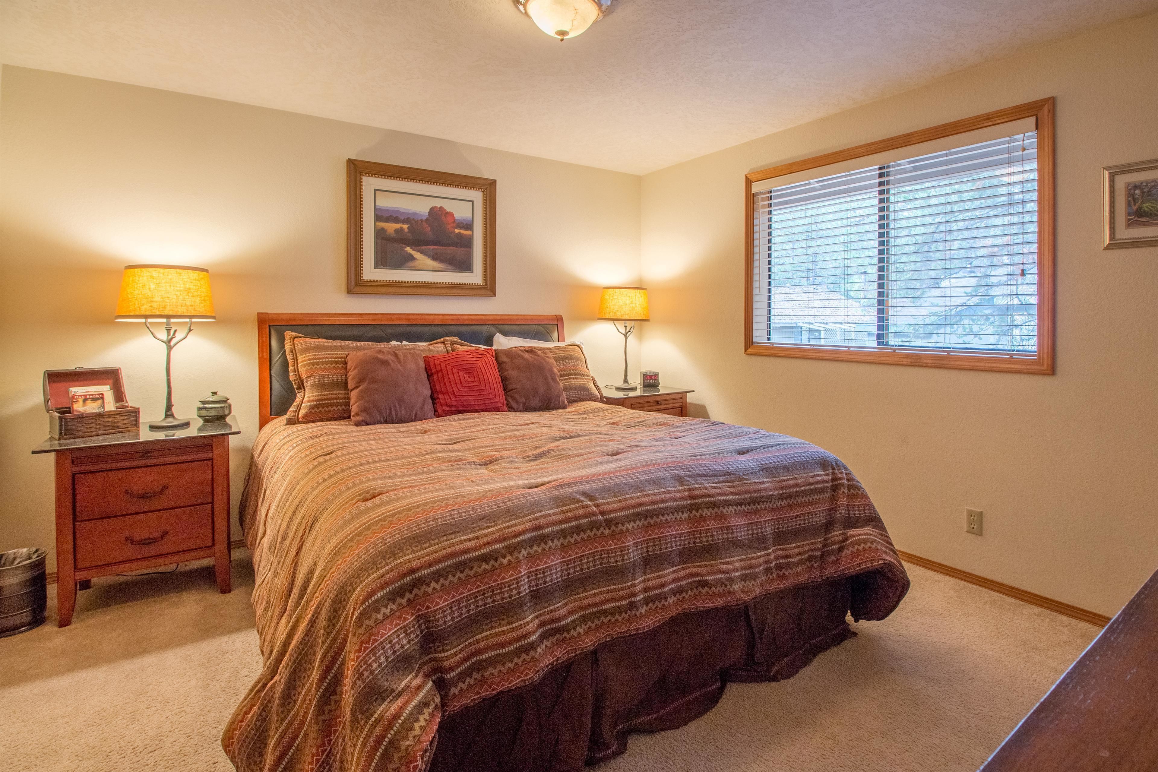A bedroom with a large bed, two bedside tables with lamps, a window with blinds, and a framed picture on the wall.