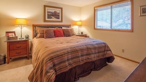 A bedroom with a large bed, two bedside tables with lamps, a window with blinds, and a framed picture on the wall.